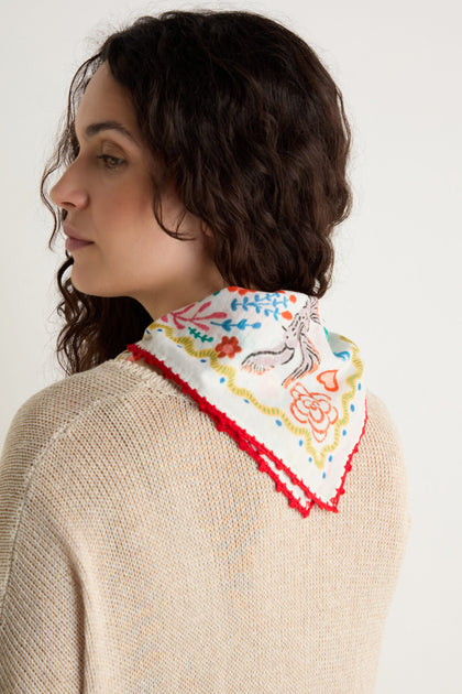 A woman with wavy dark hair wears a cream knit sweater and drapes the Embroidered Love In Bloom Cotton Bandana, featuring colorful floral patterns and red trim, over her shoulders.