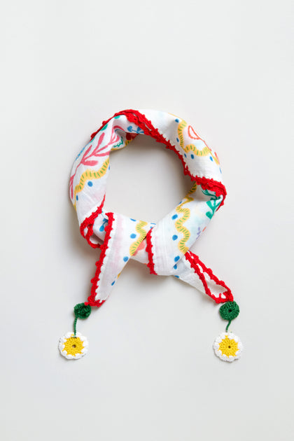 The Embroidered Love In Bloom Cotton Bandana features colorful abstract patterns, red trim, and crocheted daisy flowers at both ends, all on white cotton. Displayed on a light background.