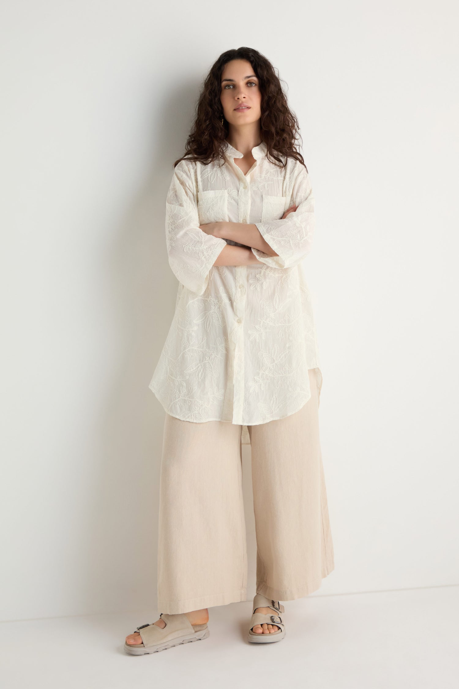 A woman stands against a white wall wearing an Embroidered Long Floral Shirt, wide beige pants, and beige sandals.