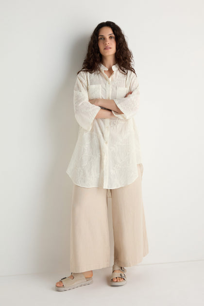 A woman stands against a white wall wearing an Embroidered Long Floral Shirt, wide beige pants, and beige sandals.