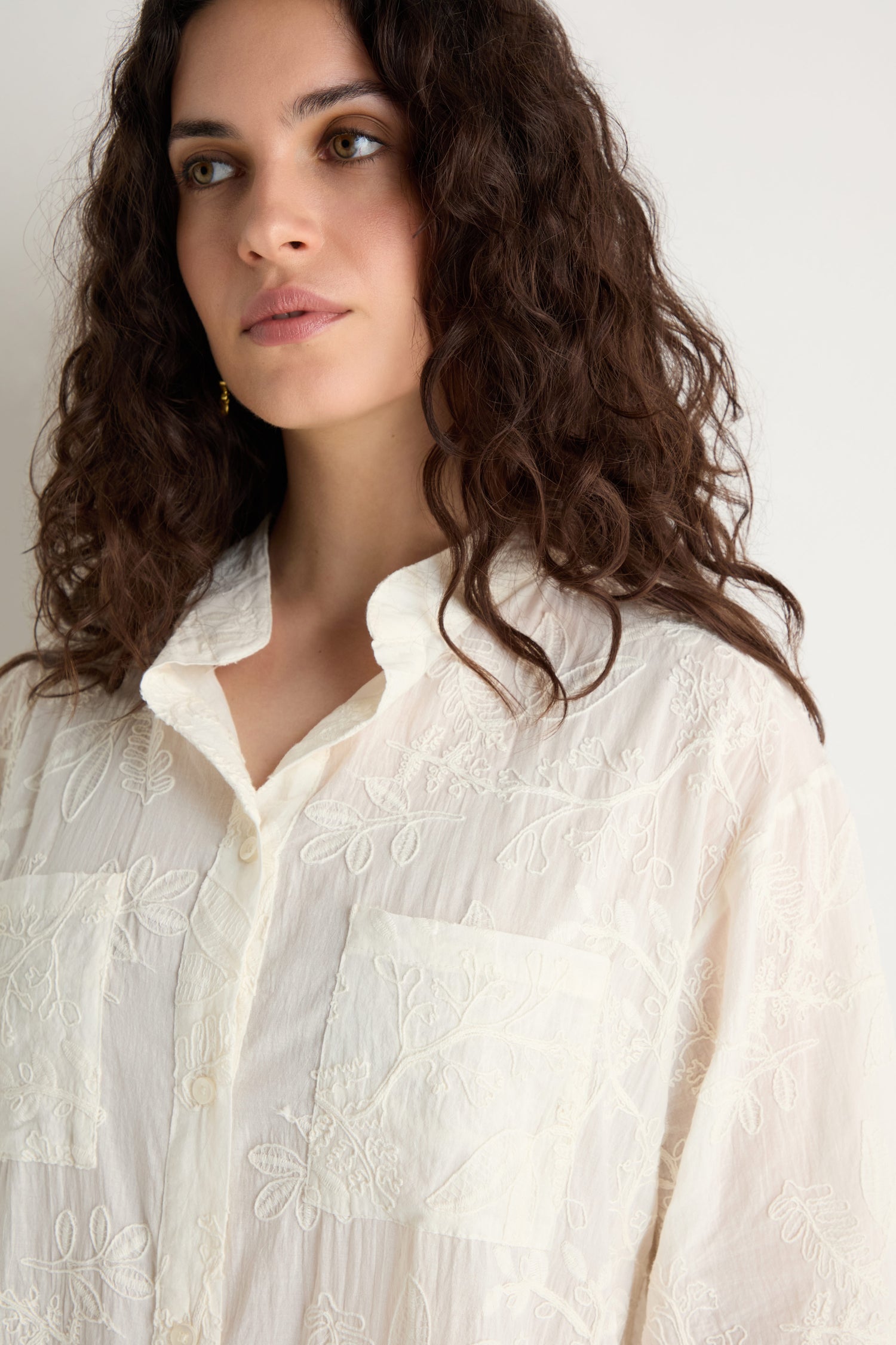 A woman with wavy brown hair wears the Embroidered Long Floral Shirt, featuring a white fabric, floral embroidery, and two front pockets, standing against a plain light background.