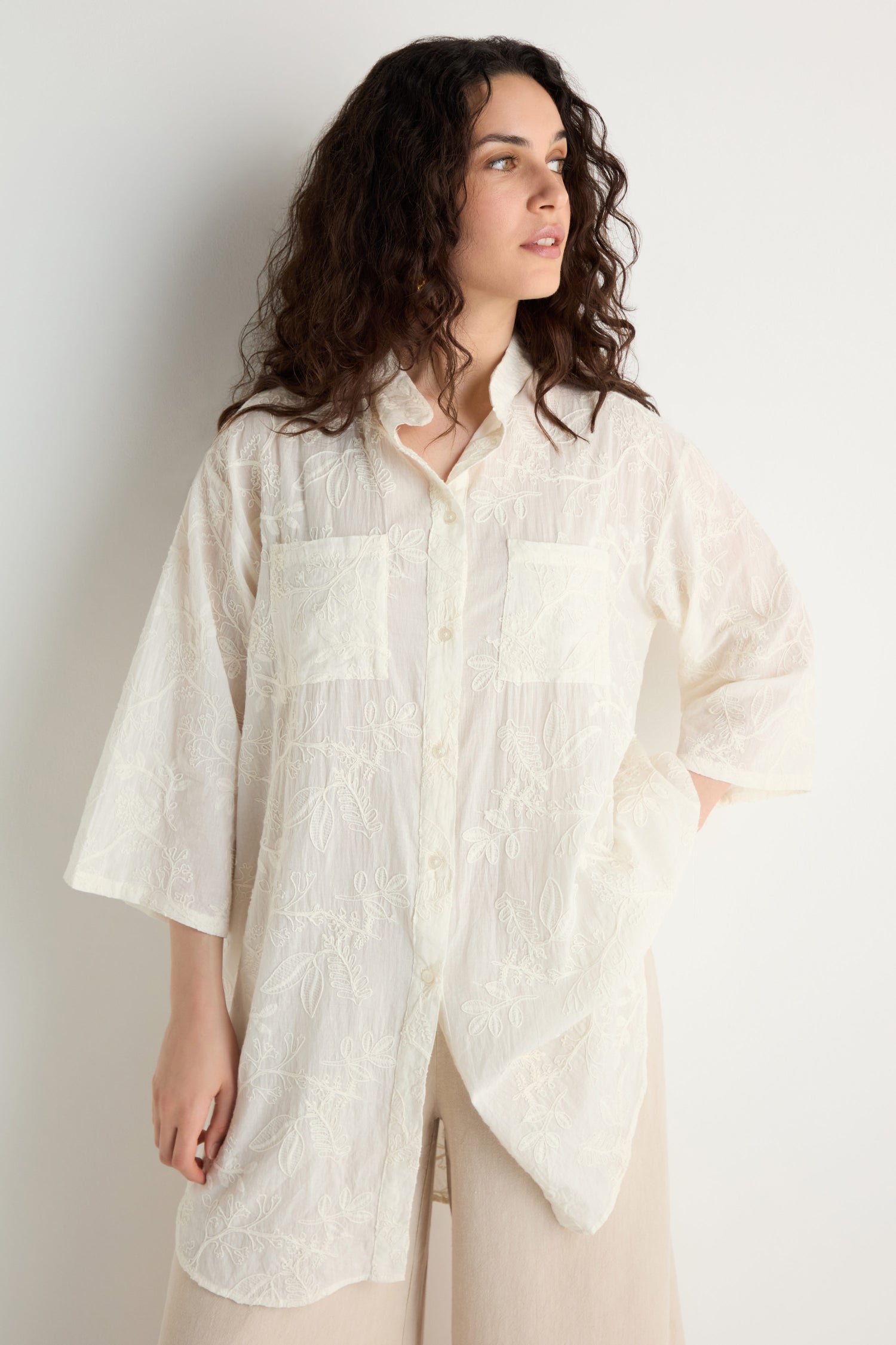 Woman with wavy dark hair wears the Embroidered Long Floral Shirt, a loose, semi-sheer top with floral embroidery and two front pockets, posing against a plain light background.
