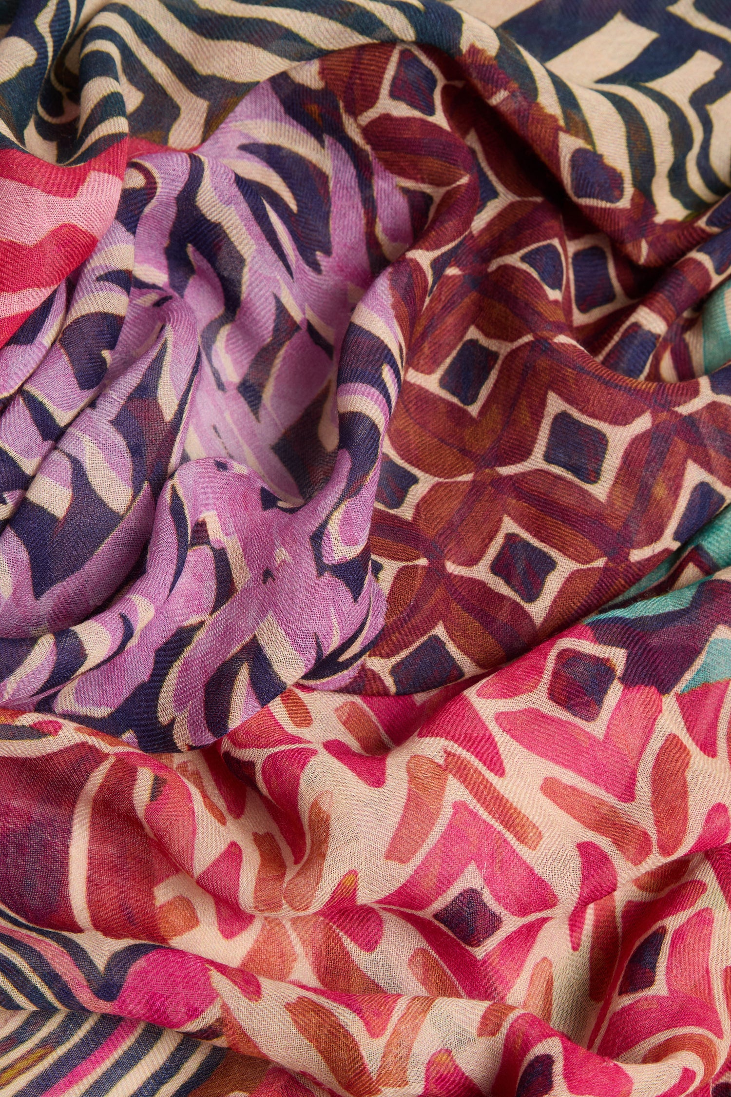 A close-up of the Graphic Print Wool Scarf, featuring a vibrant patchwork of abstract geometric and leaf motifs in shades of purple, red, brown, pink, green, and cream. The wool textile is softly draped and layered to showcase its striking graphic print design.