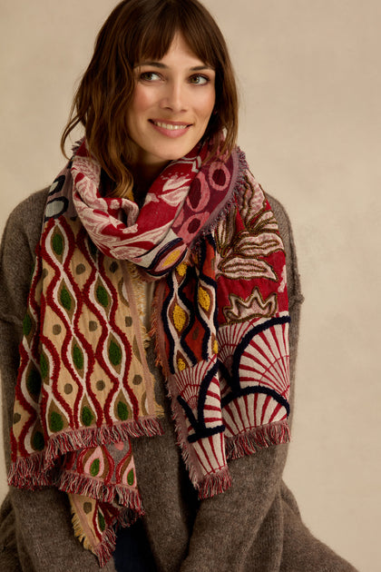 A woman smiles at the camera against a neutral background, wearing a brown sweater and the Embroidered Cotton Wool Scarf, which is adorned with vibrant red, green, and cream geometric and floral motifs. The scarf features delicate fringed edges for an added touch of elegance.