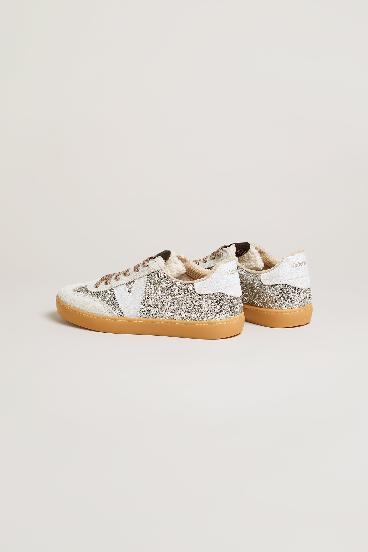 The Berlin Glitter Trainers are a pair of sparkly silver sneakers with white accents, tan soles, and brown laces, highlighted by stylish suede panels and displayed against a plain light background.
