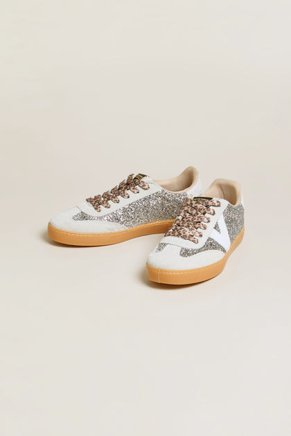 The Berlin Glitter Trainers are a pair of low-top sneakers featuring silver glitter details, white accents, tan soles, brown laces, and stylish suede panels, presented against a plain light background.