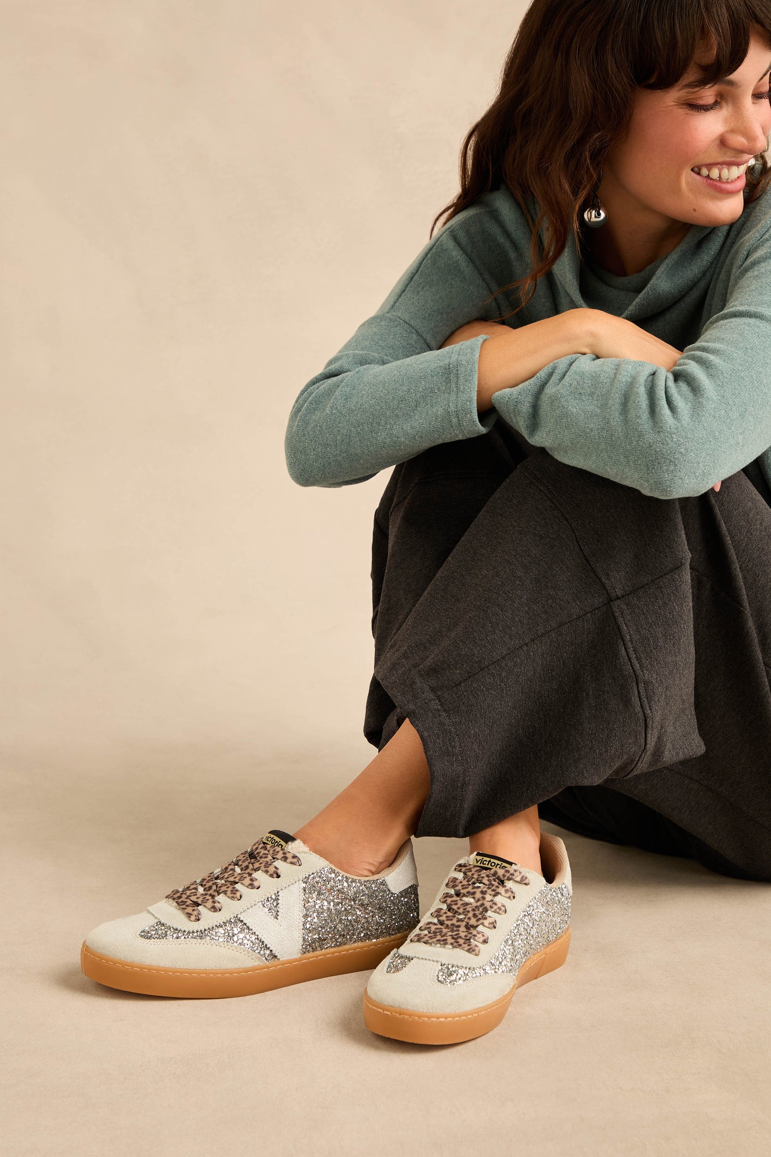 A person in a teal sweater and dark pants sits on the floor, smiling, wearing Berlin Glitter Trainers featuring silver uppers, white accents, brown soles, suede panels, and leopard print laces.