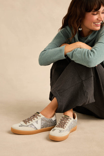 A person in a teal sweater and dark pants sits on the floor, smiling, wearing Berlin Glitter Trainers featuring silver uppers, white accents, brown soles, suede panels, and leopard print laces.