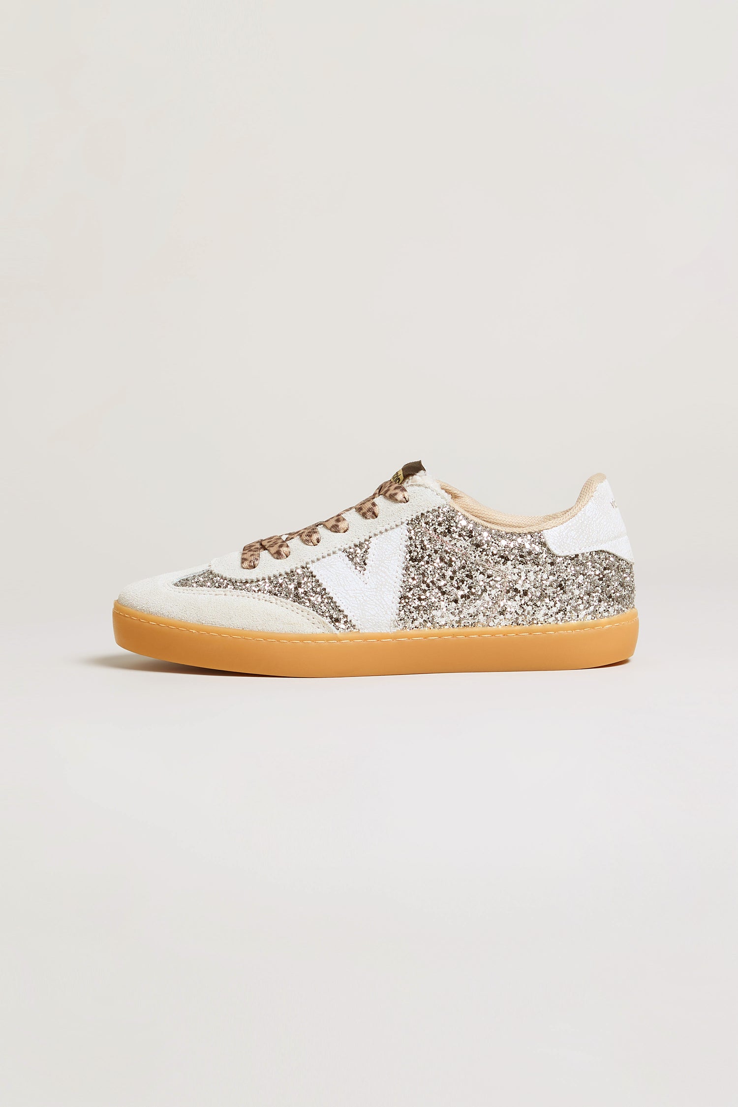 Showcasing the Berlin Glitter Trainers, this low-top design features striking white and silver glitter panels complemented by eye-catching suede accents. The trainers are finished with brown laces, a white side logo, a light brown rubber sole, and an animal-print heel accent, all set against a plain light background.