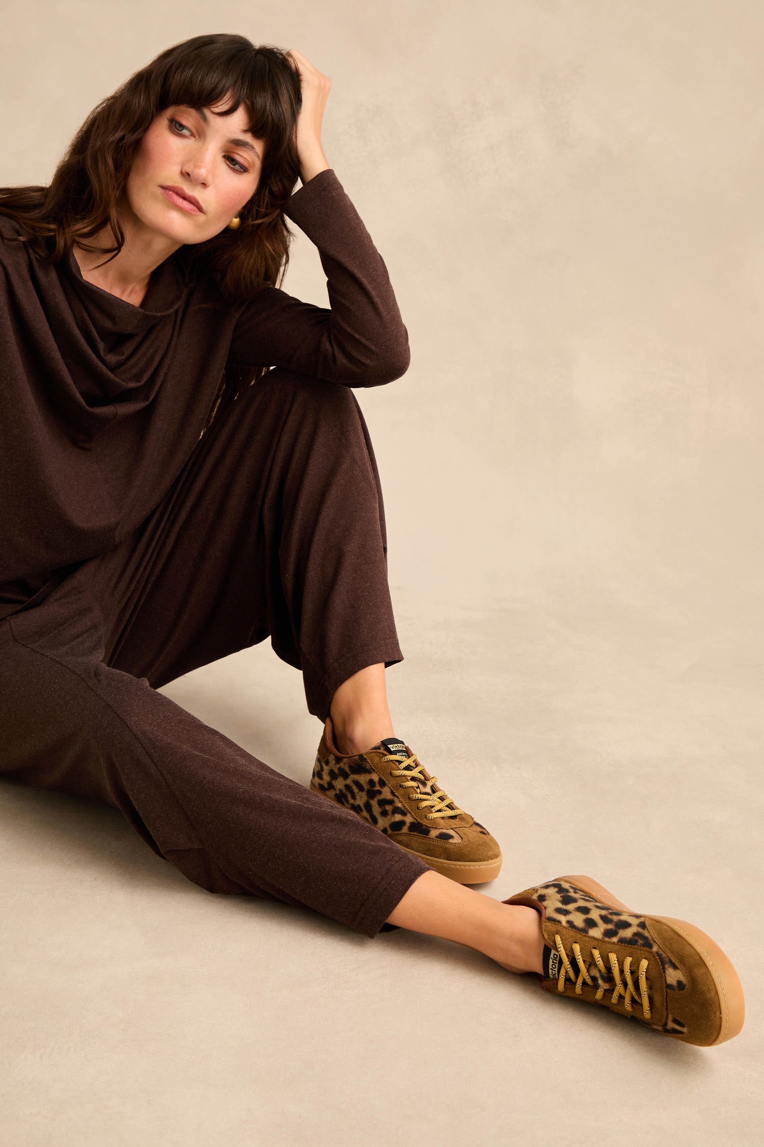 A woman in a brown outfit sits on the floor with one arm resting on her knee, wearing Berlin Animal Print Trainers made in Spain, and looks slightly upward.