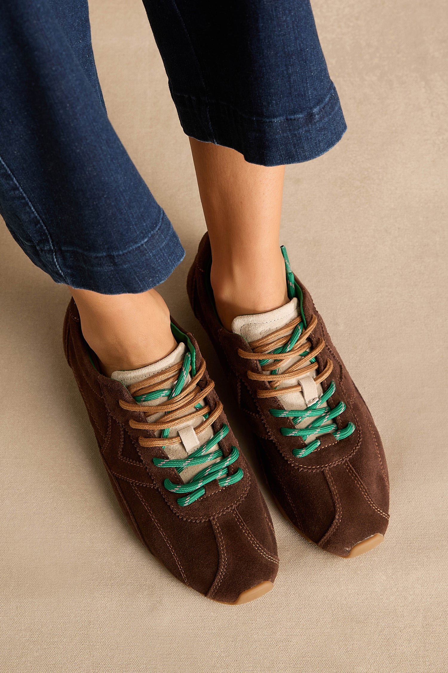 A person wearing the Aster Suede Trainers in dark brown cowhide with green and tan laces and subtle contrast stitching, paired with cropped dark blue jeans, stands on a light beige surface.