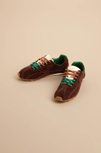 A pair of Aster Suede Trainers in brown cowhide suede featuring green inner lining, beige soles, contrast stitching, and mismatched beige and green laces, displayed on a beige background.