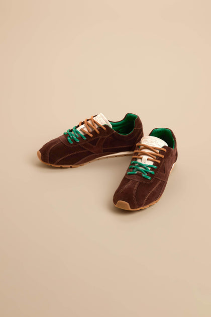 A pair of Aster Suede Trainers in brown cowhide suede featuring green inner lining, beige soles, contrast stitching, and mismatched beige and green laces, displayed on a beige background.