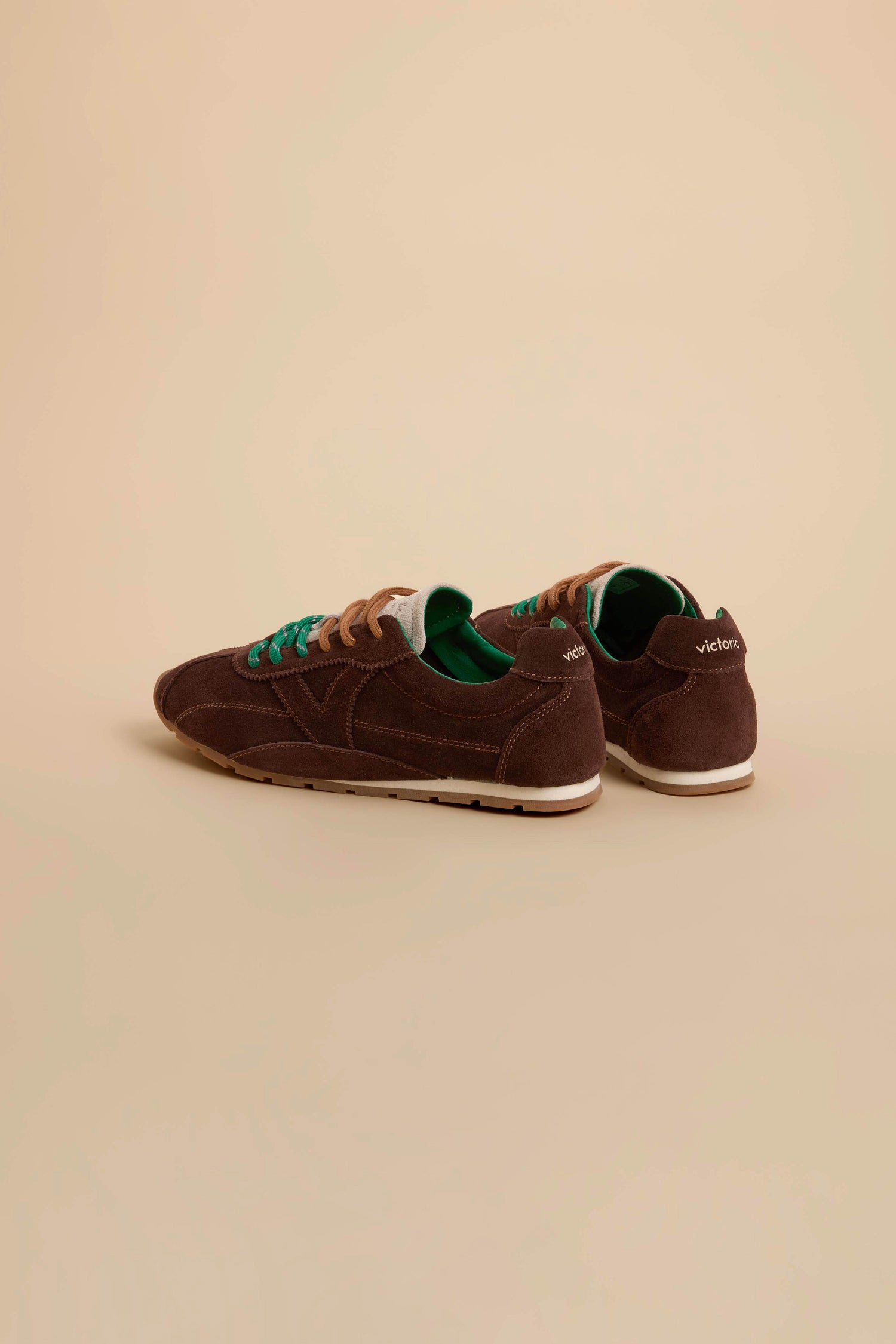A pair of Aster Suede Trainers in brown cowhide suede with tan soles, teal laces, and green inner lining, featuring subtle contrast stitching, displayed on a beige background.