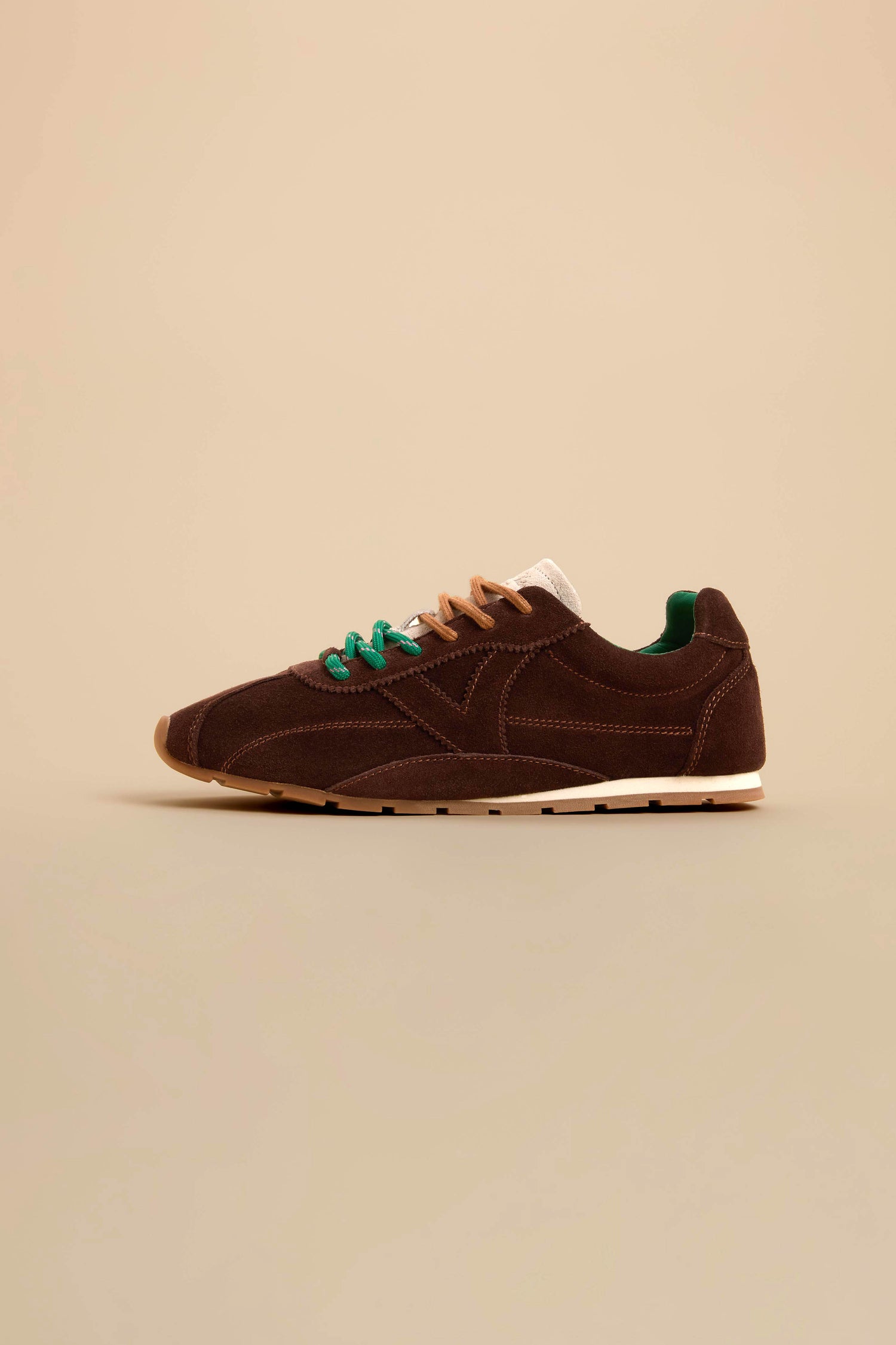 Aster Suede Trainers in brown cowhide suede feature a white sole, tan and green laces, contrast stitching, and green accents, displayed in profile against a beige background.