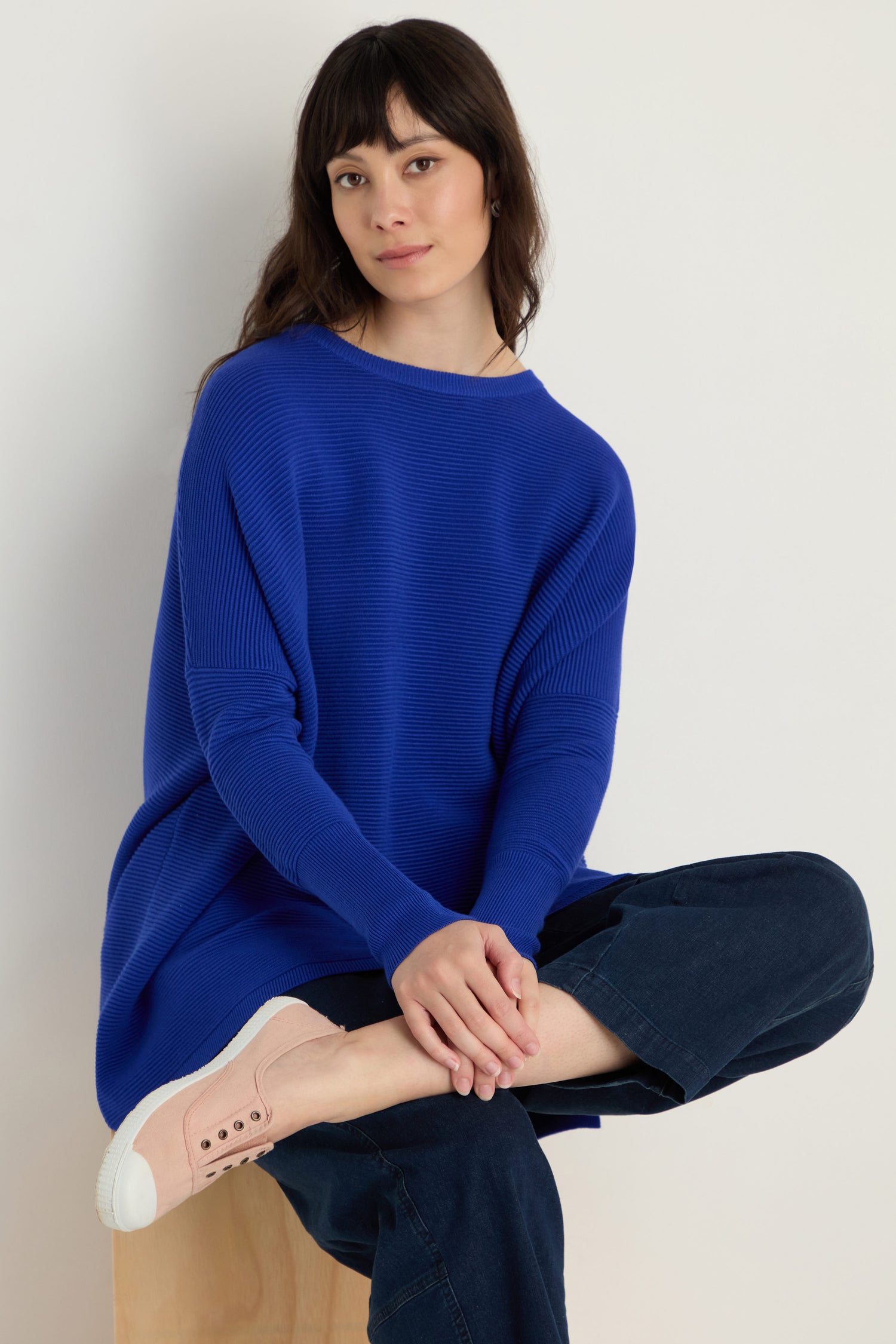 A woman sits on a wooden stool against a plain background, wearing a bright blue sweater, dark pants, and Inglesa Light Pink Pumps.