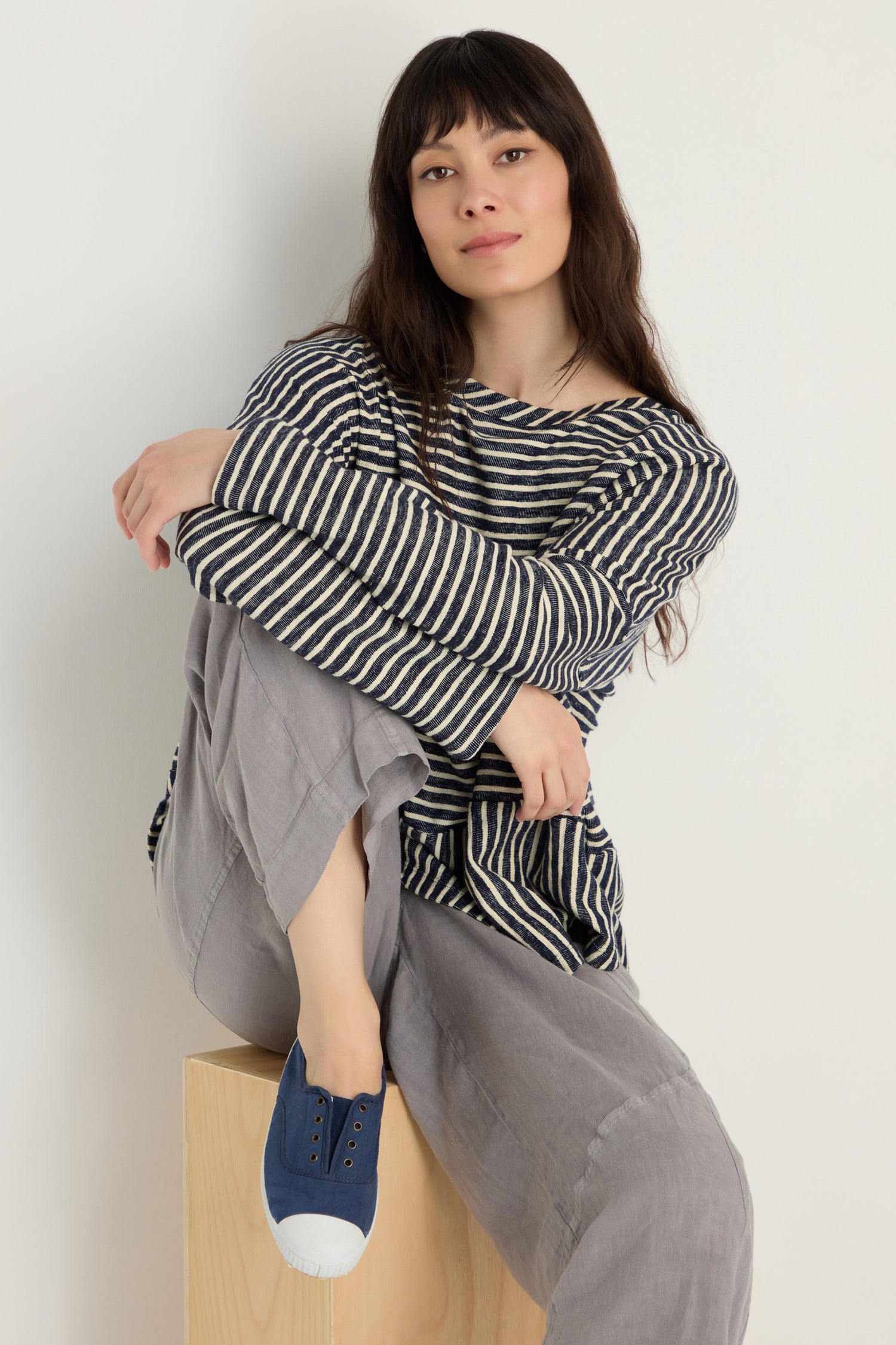 A woman with long brown hair sits on a wooden block, wearing a striped long-sleeve shirt, light gray pants, and Inglesa Navy Pumps made in Spain, looking at the camera.