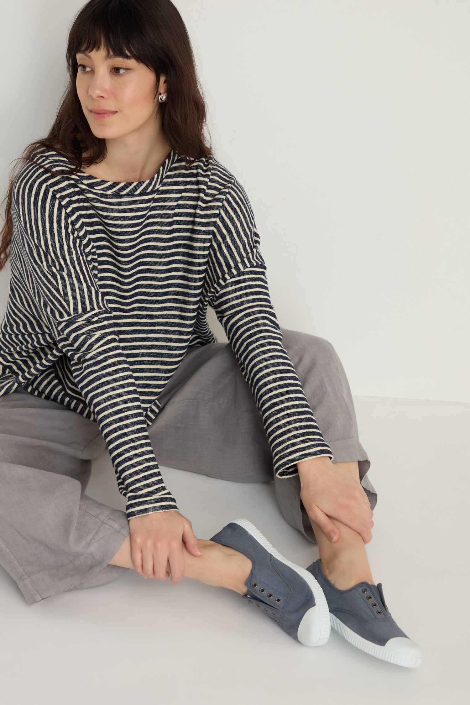 A woman in a black and white striped long-sleeve shirt and gray pants sits on the floor, putting on Inglesa Slate Pumps.