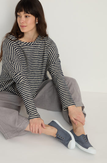 A woman in a black and white striped long-sleeve shirt and gray pants sits on the floor, putting on Inglesa Slate Pumps.