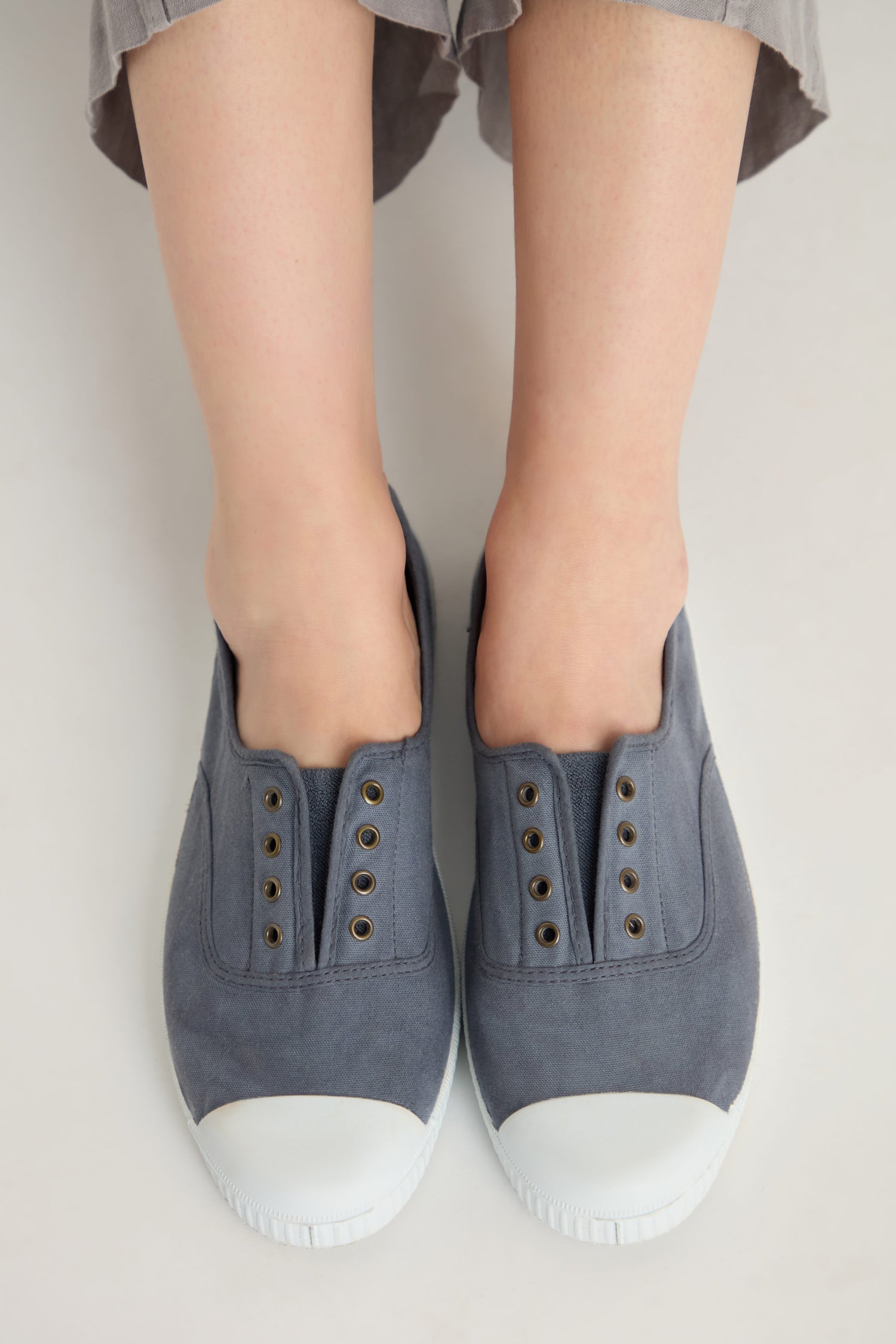 A person wearing Inglesa Slate Pumps—gray slip-on canvas trainers with white rubber toes, made in Spain from recycled cotton and featuring a laceless design—stands on a light-colored surface.