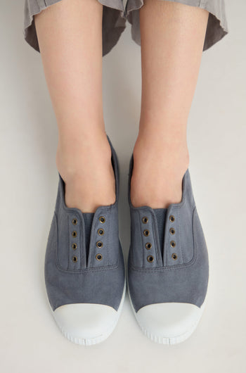 A person wearing Inglesa Slate Pumps—gray slip-on canvas trainers with white rubber toes, made in Spain from recycled cotton and featuring a laceless design—stands on a light-colored surface.