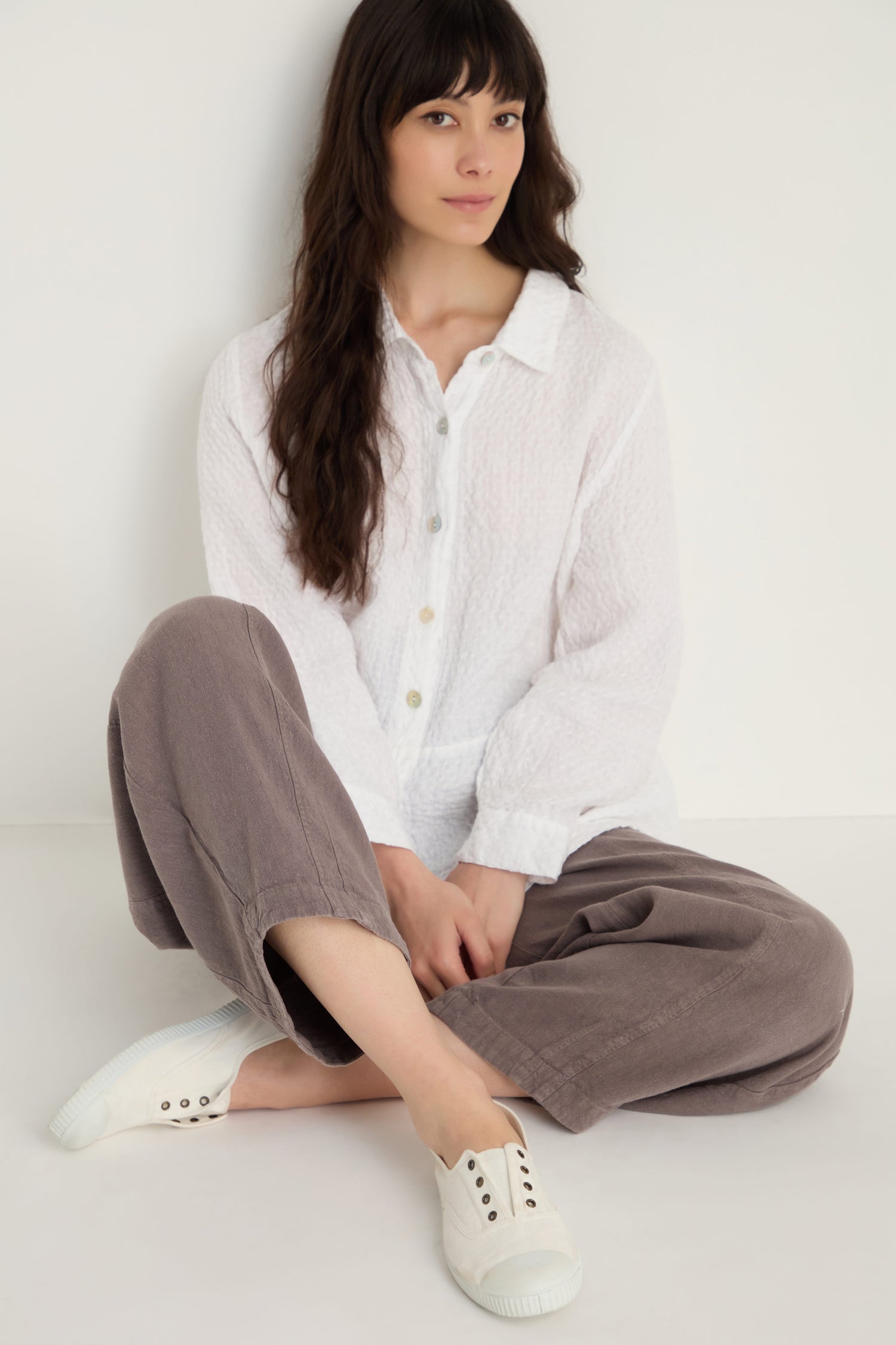 A person with long dark hair sits on the floor against a white wall, wearing a textured white button-up shirt, loose taupe pants, and Inglesa White Pumps.