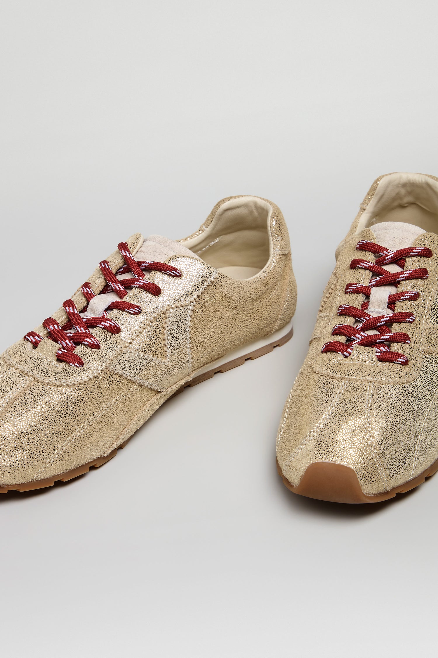 Aster Leather Trainers made from metallic gold cowhide, featuring red and white striped laces, displayed on a light gray background.