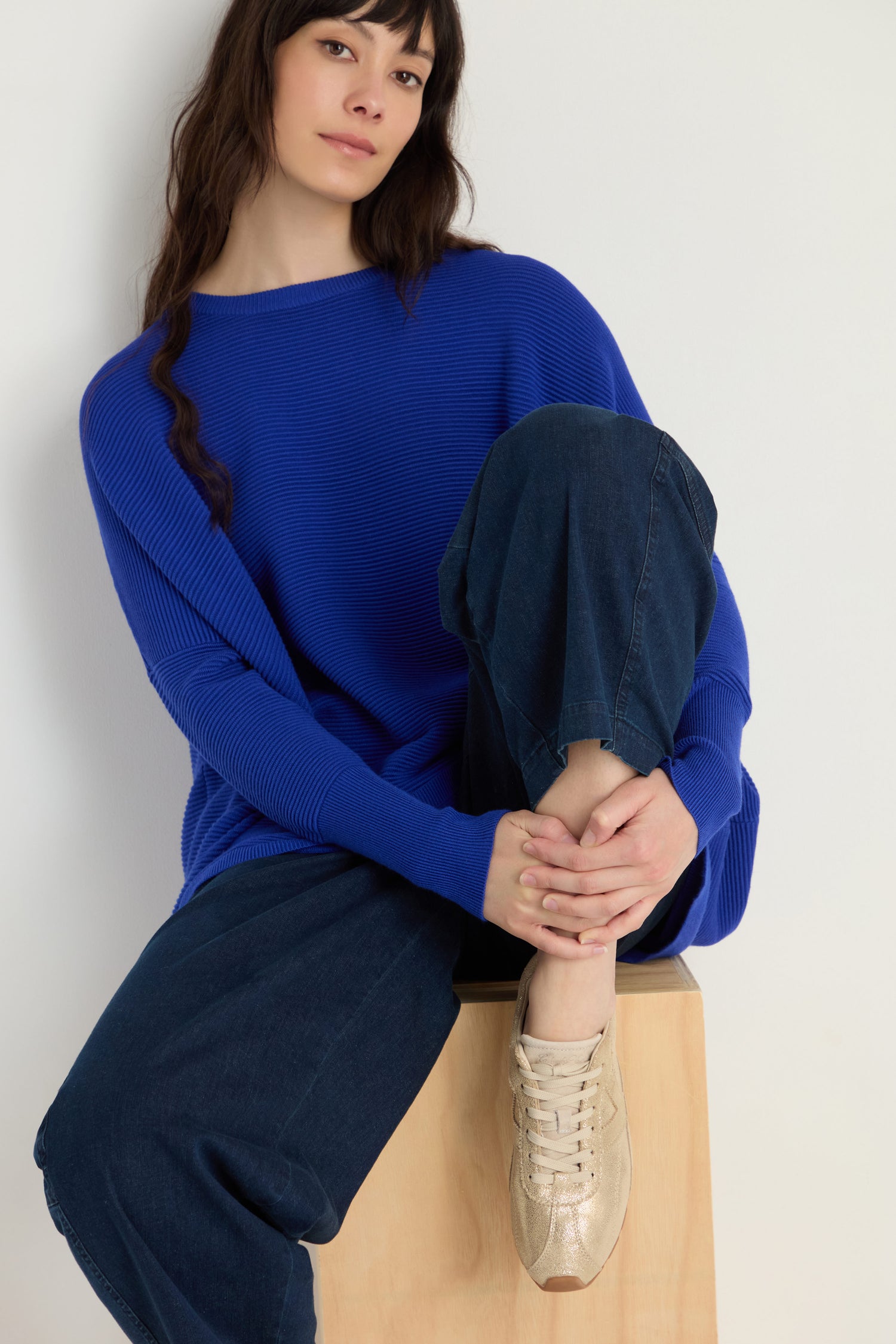 A woman in a blue sweater and wide-leg jeans sits on a wooden block, resting one foot—wearing Aster Leather Trainers—on the block and clasping her knee with both hands.
