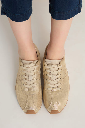 A person wearing cuffed blue jeans and Aster Leather Trainers in metallic gold cowhide stands on a light-colored surface.