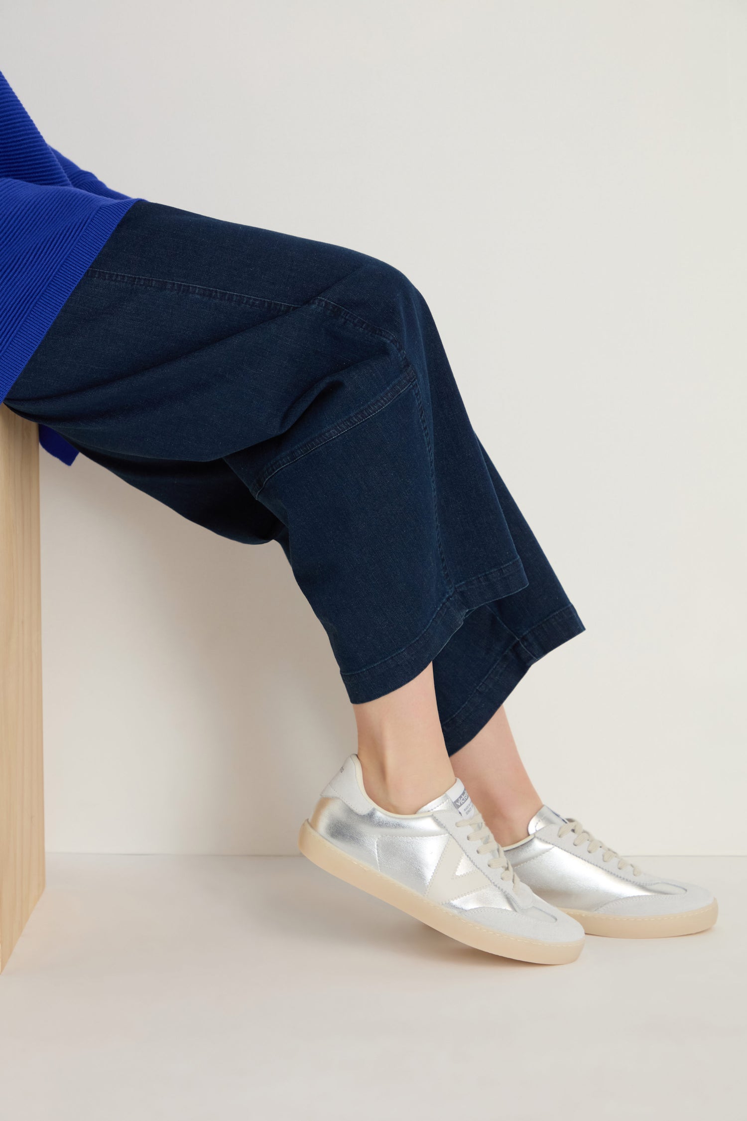 A person wears dark wide-leg jeans and Berlin Metallic Trainers with ergonomic insoles and beige soles, sitting on a light wooden bench against a plain background.
