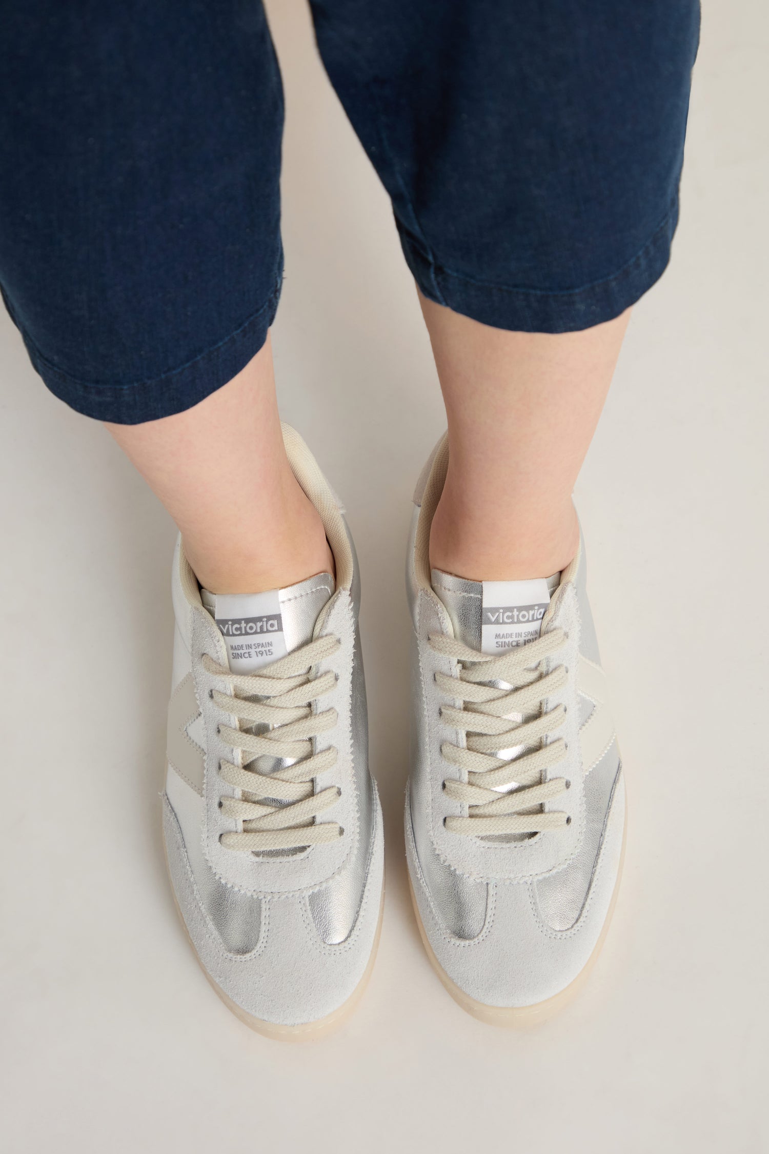 Person wearing dark blue pants and Berlin Metallic Trainers with beige laces, viewed from above on a light-colored floor.