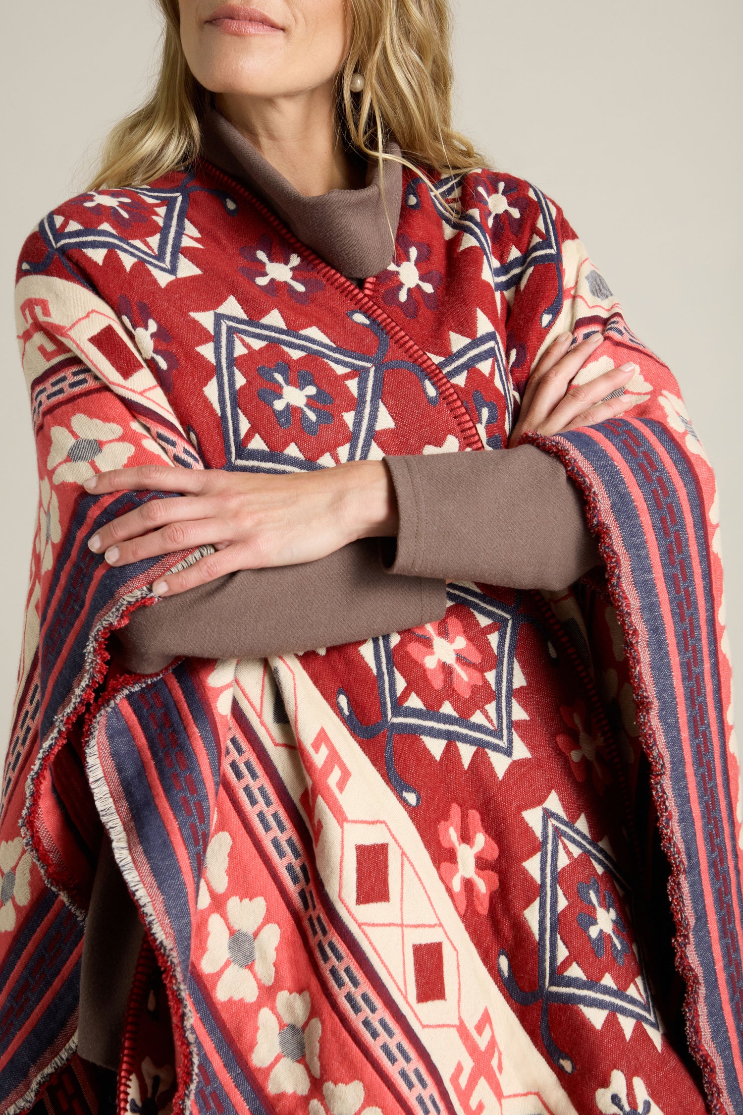 A person wearing an Aztec Woven Shawl featuring a vibrant mix of red, blue, and white with folded arms. The shawl boasts intricate geometric and floral designs, making it perfect for an autumn wardrobe.