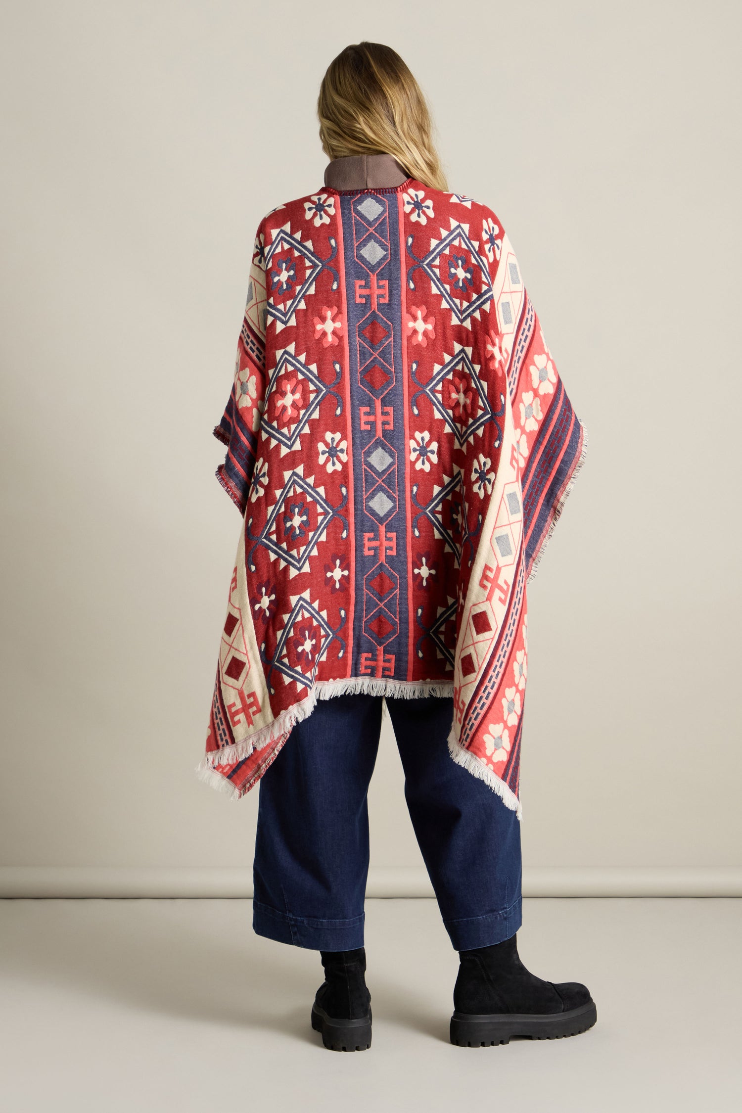 Person standing with their back to the camera, wearing the Aztec Woven Shawl with fringe over navy blue pants and black boots, perfect for an autumn wardrobe.