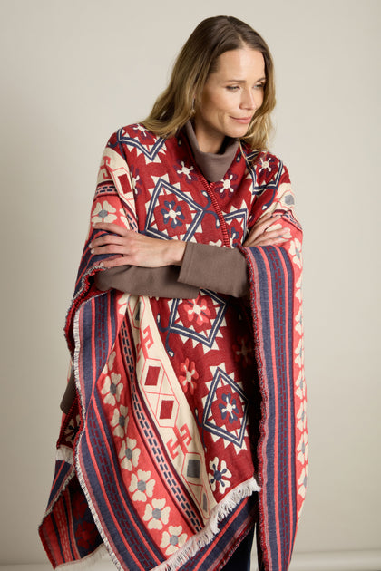 A person wrapped in the Aztec Woven Shawl gazes to the side with arms crossed. The shawl features red, blue, and white geometric designs. Perfect for an autumn wardrobe, this shawl adds a touch of warmth and style.