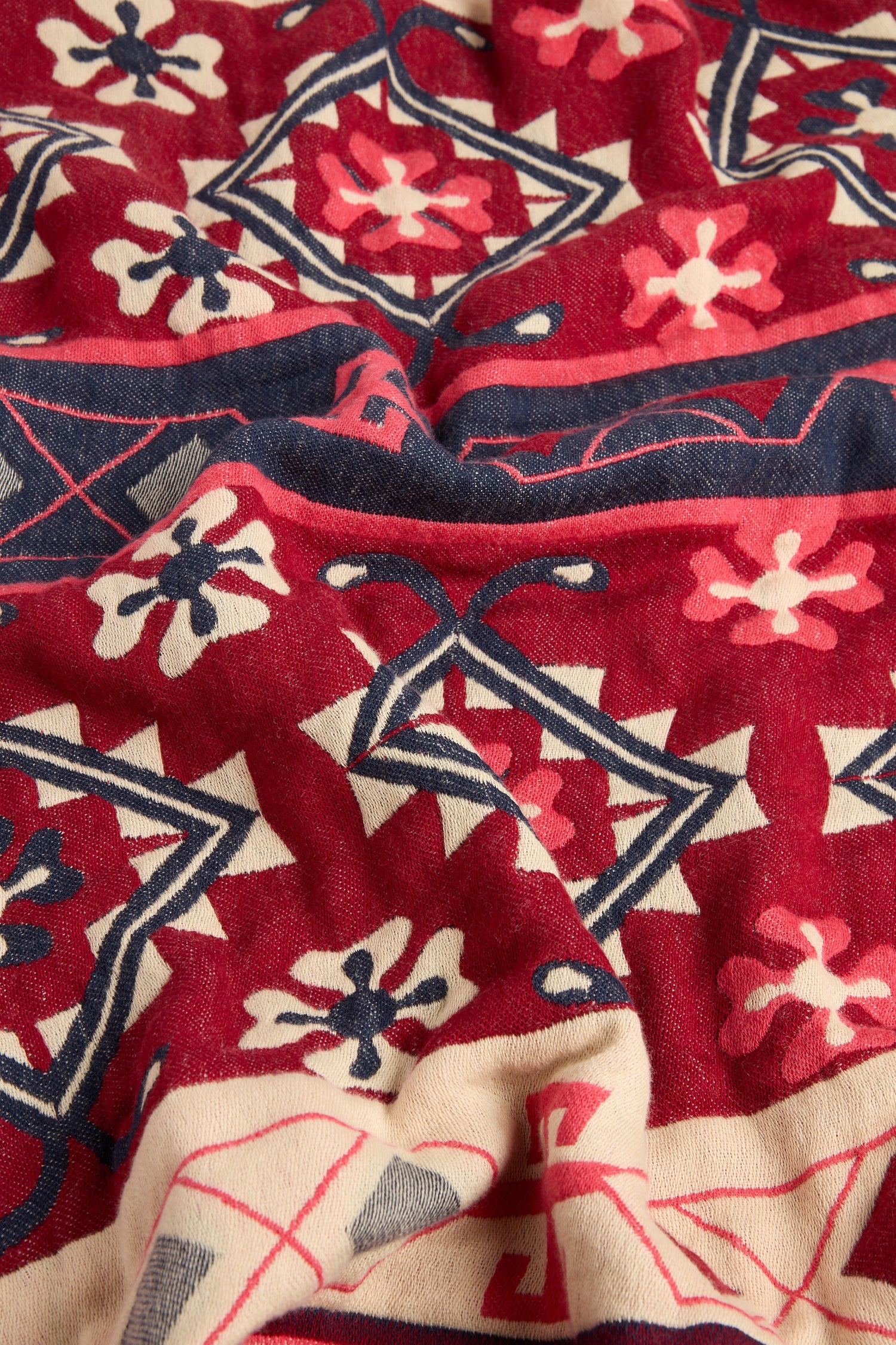 A close-up view of a fabric with intricate geometric and floral patterns in shades of red, blue, pink, and cream. The fabric has a soft texture and slight wrinkles, reminiscent of the Aztec Woven Shawl that is perfect for an autumn wardrobe.