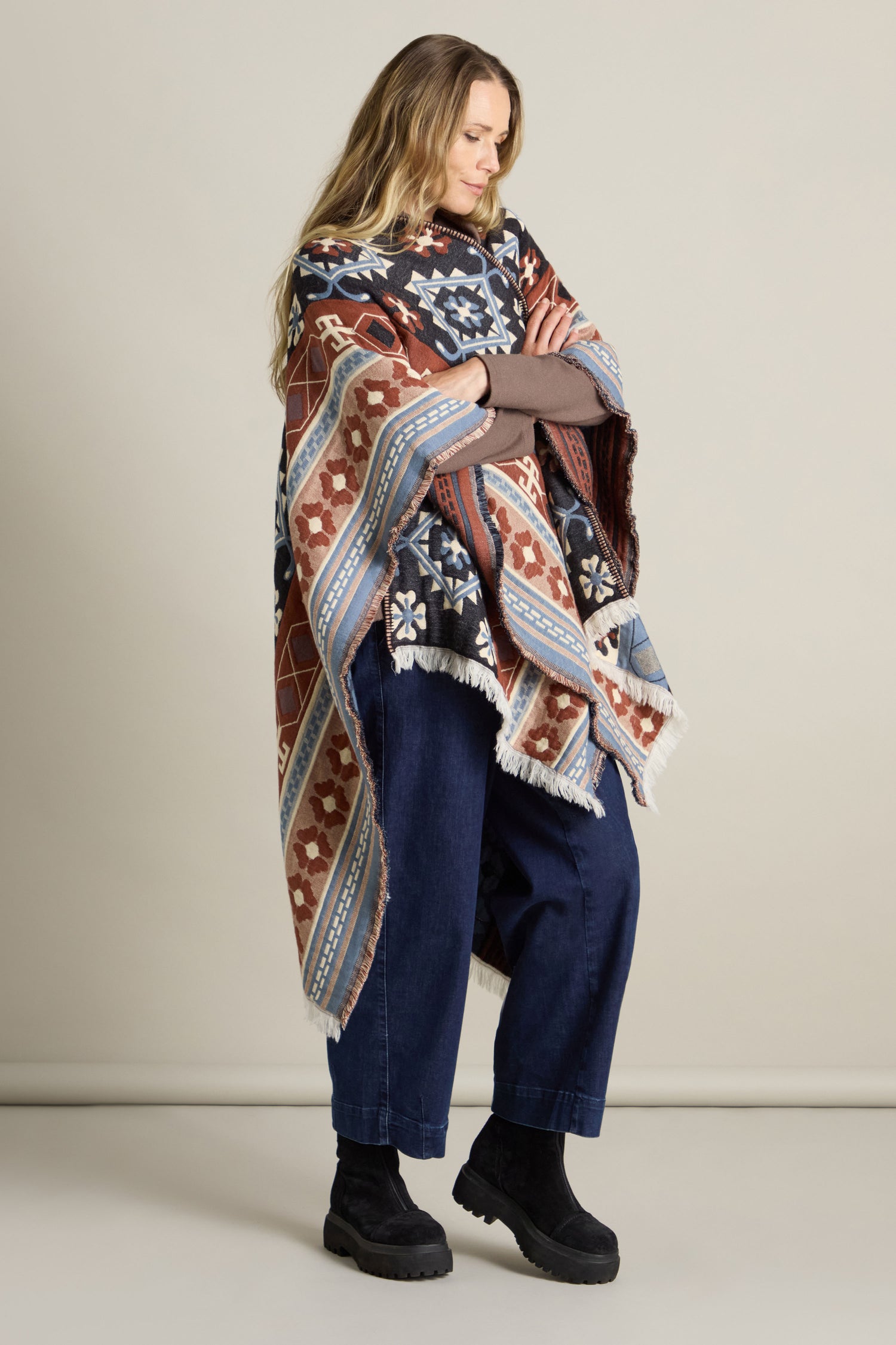 A person stands holding an Aztec Woven Shawl, its vibrant colors infusing flair into their autumn wardrobe. Dressed in a brown long-sleeve shirt, blue jeans, and black boots, they are complimented by a studio setting with a neutral background that accentuates the intricate design of the shawl.