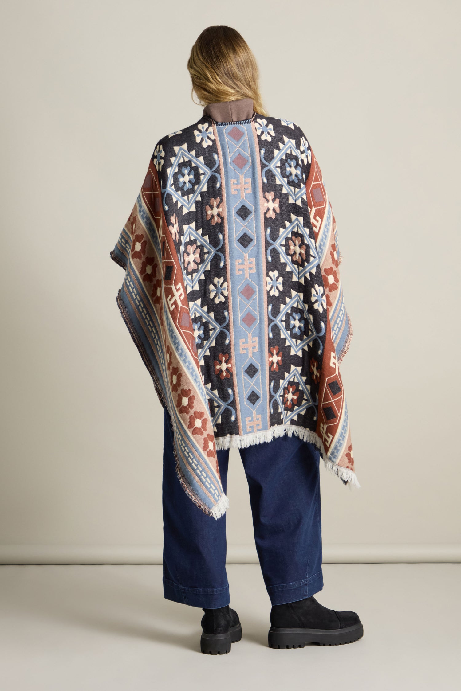 Person facing away, draped in the Aztec Woven Shawl, paired with wide-leg jeans and black boots, against a plain background—perfect for your autumn wardrobe.