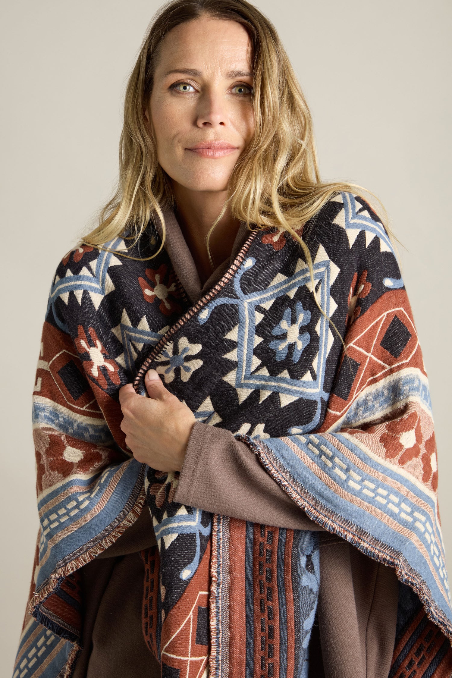 A woman with long blonde hair stands facing the camera, wrapped in the vibrant Aztec Woven Shawl adorned with a colorful geometric pattern. She has a neutral expression.