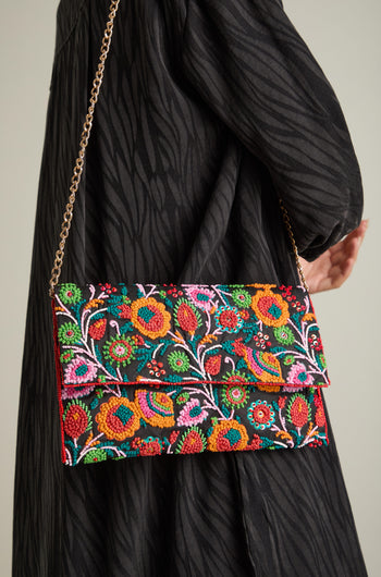 A person holds the Full Bloom Hand Beaded Clutch Bag with a gold chain strap. The clutch showcases vibrant floral beauty and is worn against a black, textured garment.