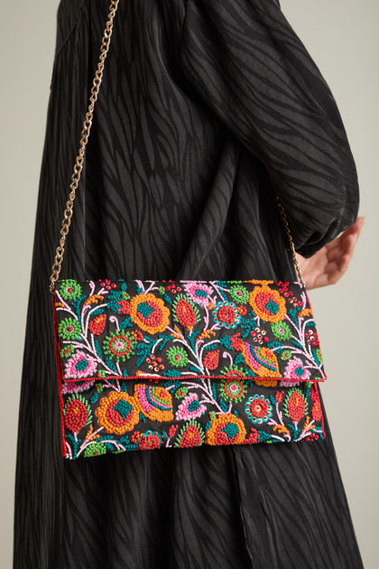 A person holds the Full Bloom Hand Beaded Clutch Bag with a gold chain strap. The clutch showcases vibrant floral beauty and is worn against a black, textured garment.