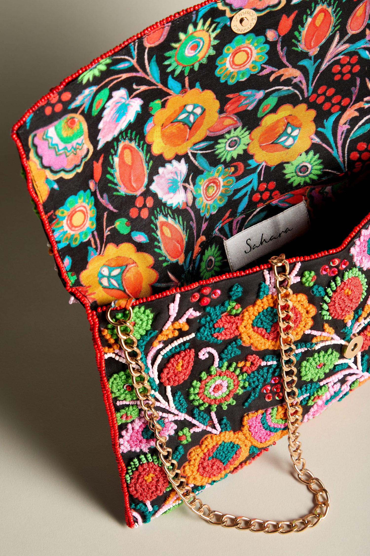 The Full Bloom Hand Beaded Clutch Bag features a floral embroidered design and a chic chain strap, and is shown open to reveal a colorful fabric lining.