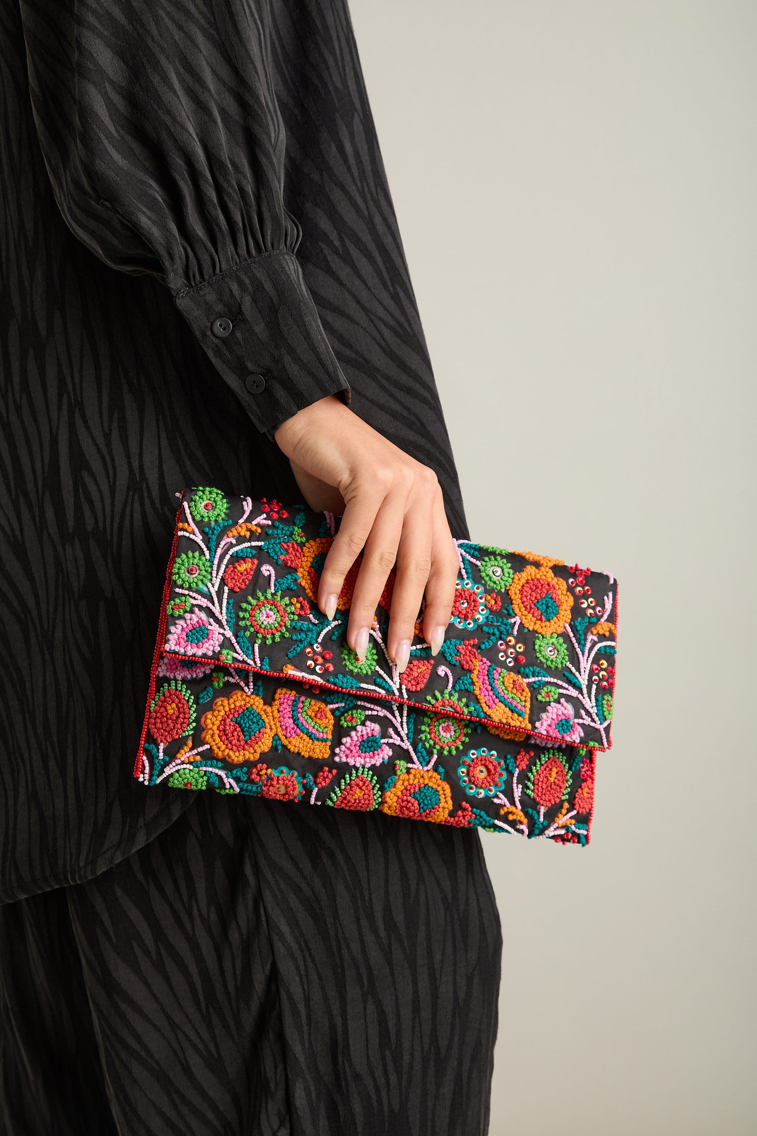 A person dressed in black holds the Full Bloom Hand Beaded Clutch Bag, adorned with a vibrant floral pattern, in their left hand.