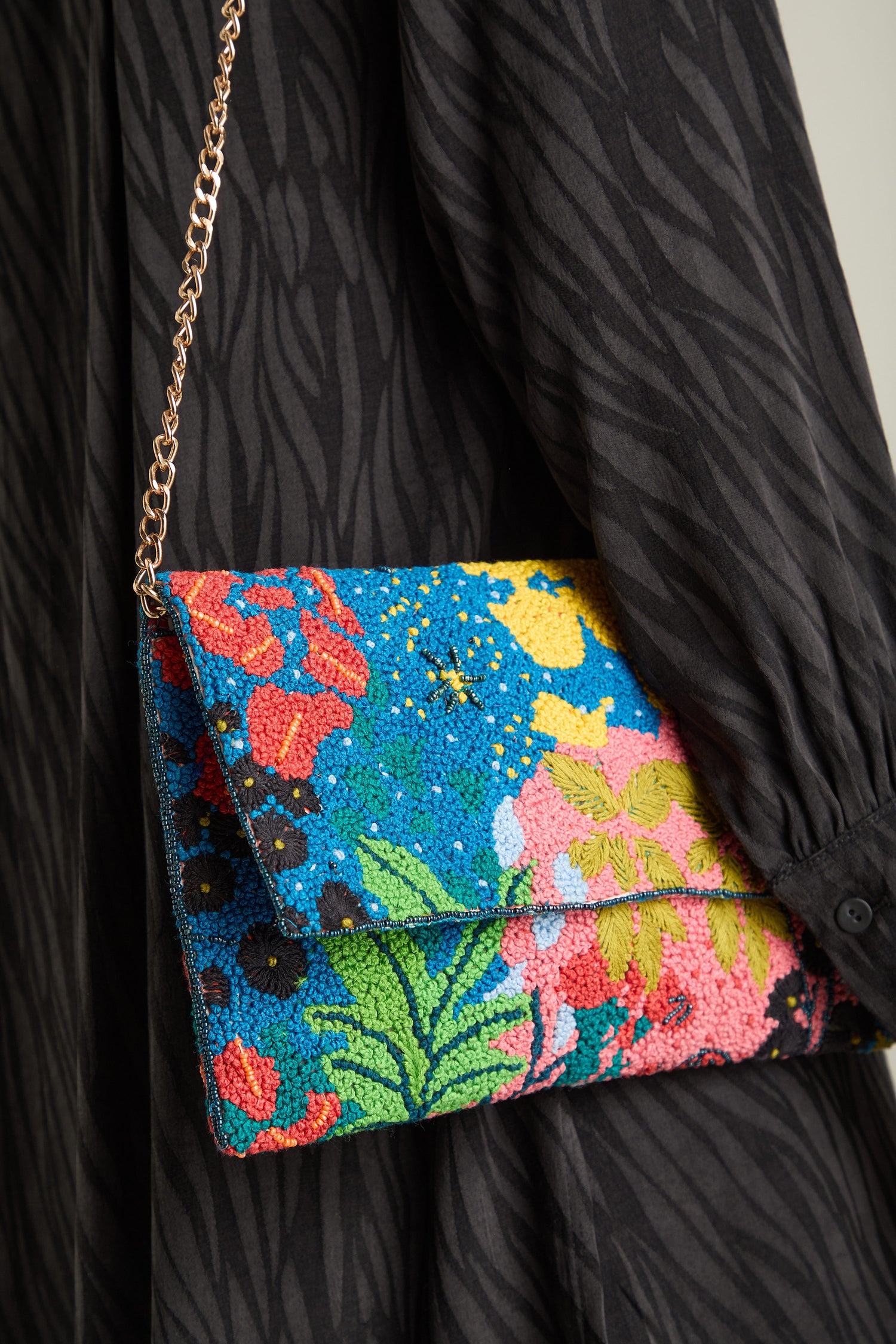 A person wearing a black patterned dress holds the Full Bloom Hand Beaded Clutch Bag, which showcases a floral beauty with its colorful beads and chic chain strap.