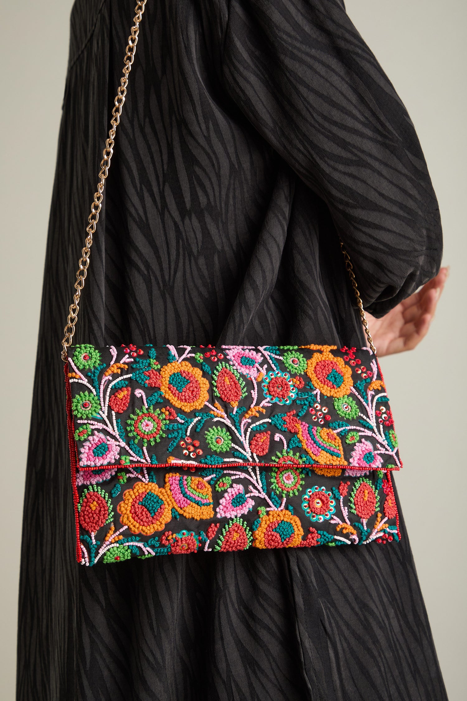 A person wearing a black patterned dress holds the Wild Garden Hand Beaded Clutch Bag, showcasing its colorful floral beaded design and gold chain strap. This statement accessory adds an elegant touch to their ensemble.