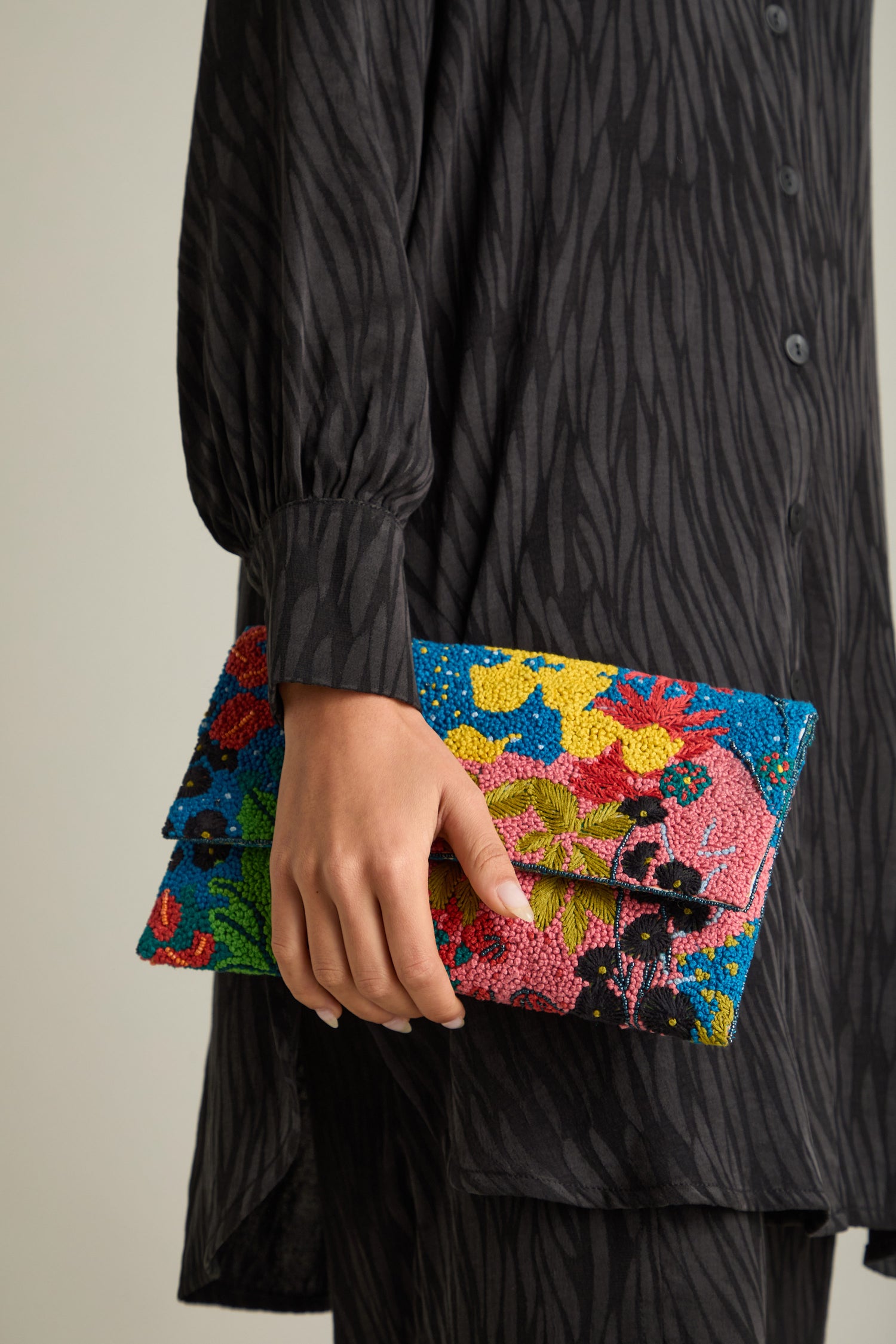 A person in a black patterned dress holds the Wild Garden Hand Beaded Clutch Bag, a vibrant statement piece featuring a stunning floral design.