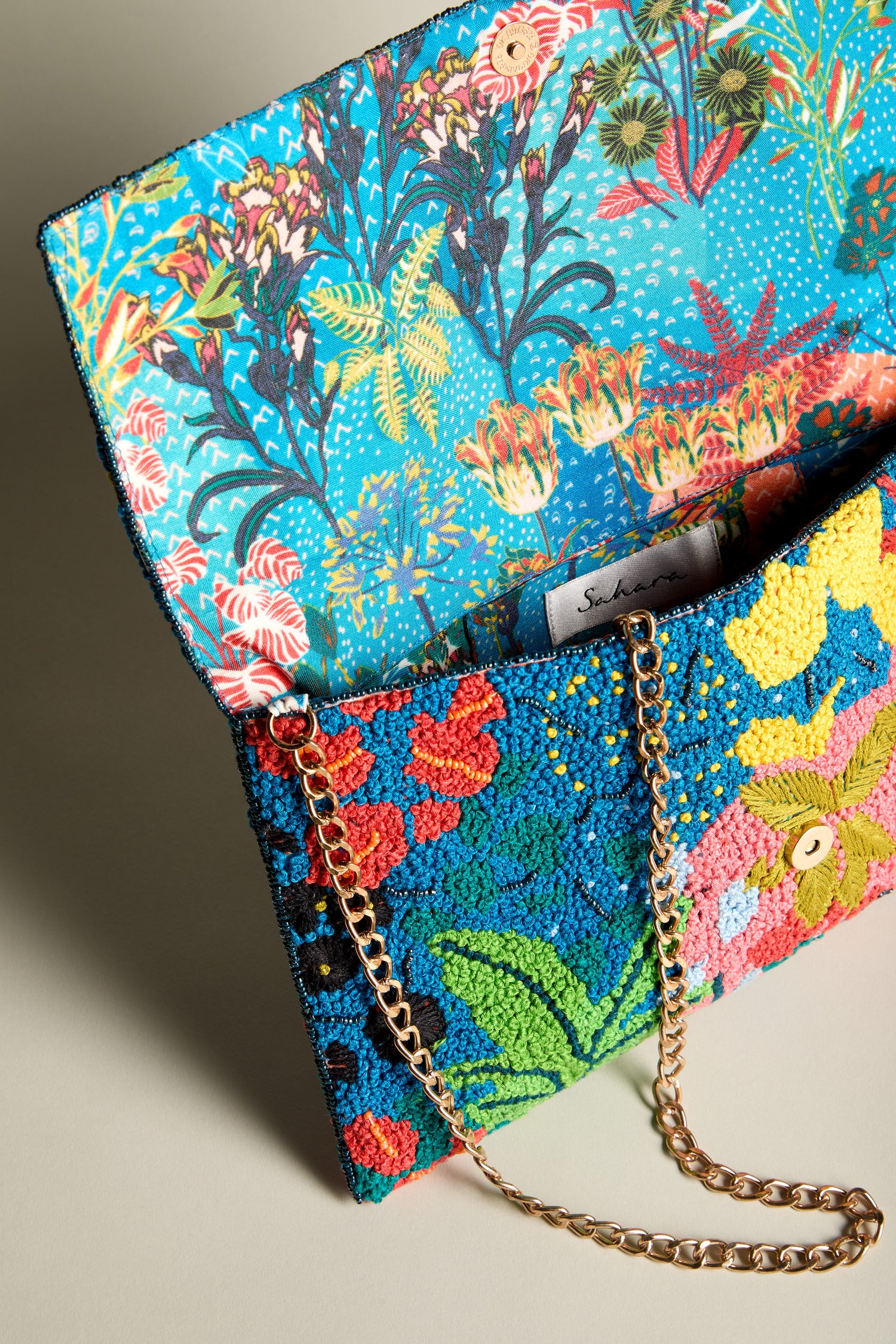 Introducing the Wild Garden Hand Beaded Clutch Bag, adorned with an exquisite floral beaded design that demands attention. This colorful clutch features a captivating floral pattern, a gold chain strap, and an intricately designed interior lining with a vibrant botanical motif. A true statement piece for any occasion.