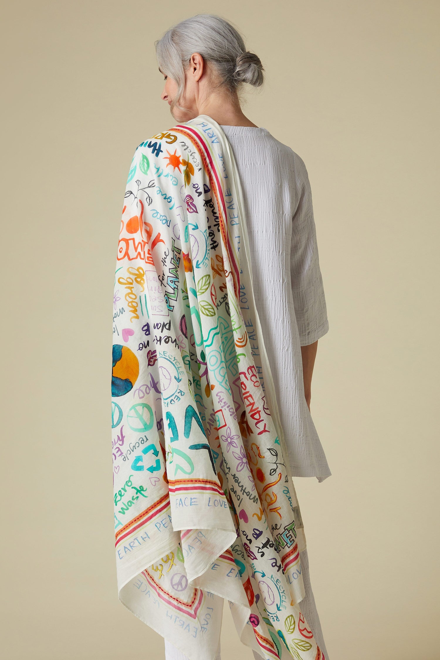 Woman in a Peace Earth Love Printed Cotton Scarf and white garment, viewed from the side against a pale background.