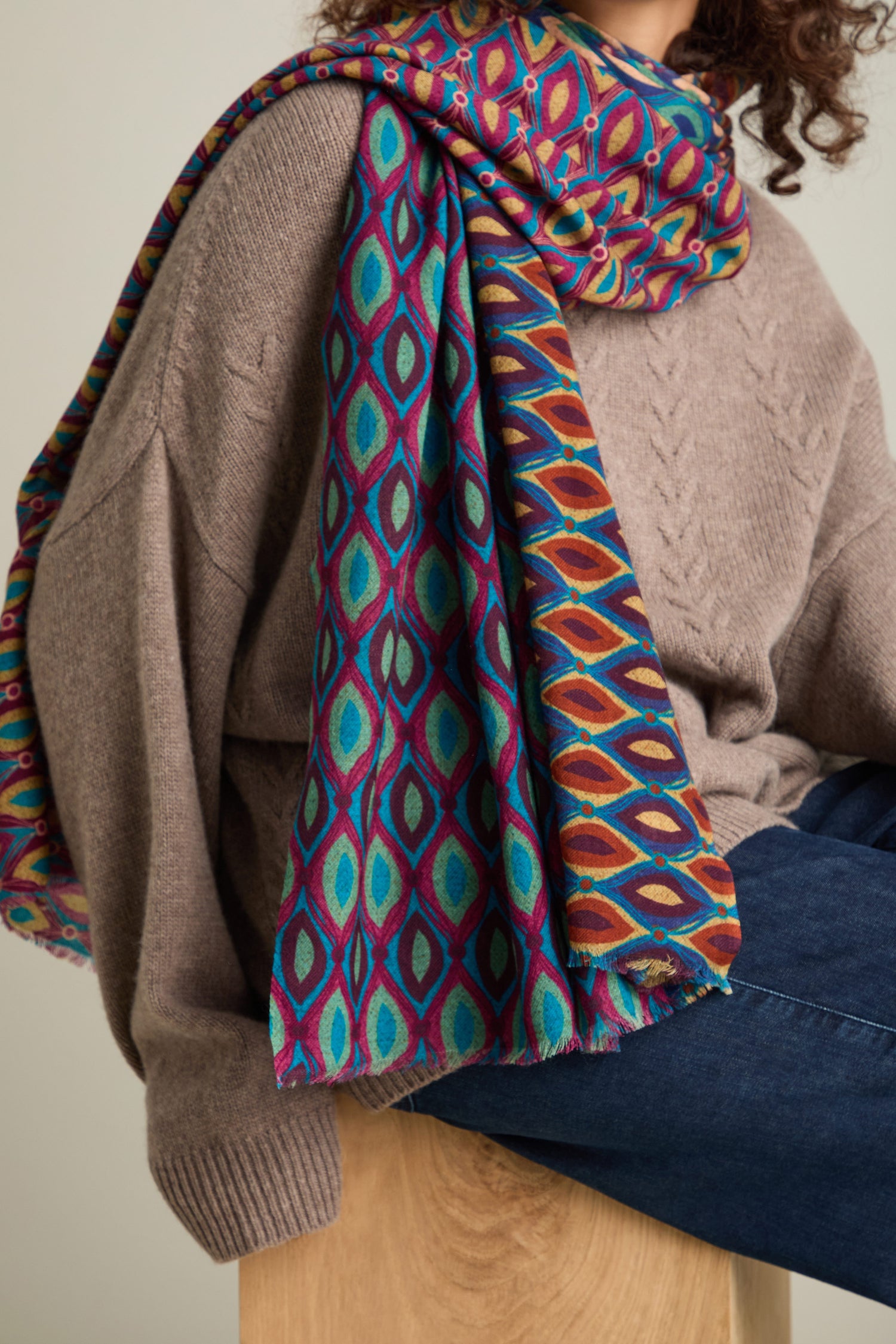 A person wearing a brown sweater and blue jeans sits with the Retro Wool Twill Scarf, featuring a colorful retro pattern print, draped around their neck and shoulder.