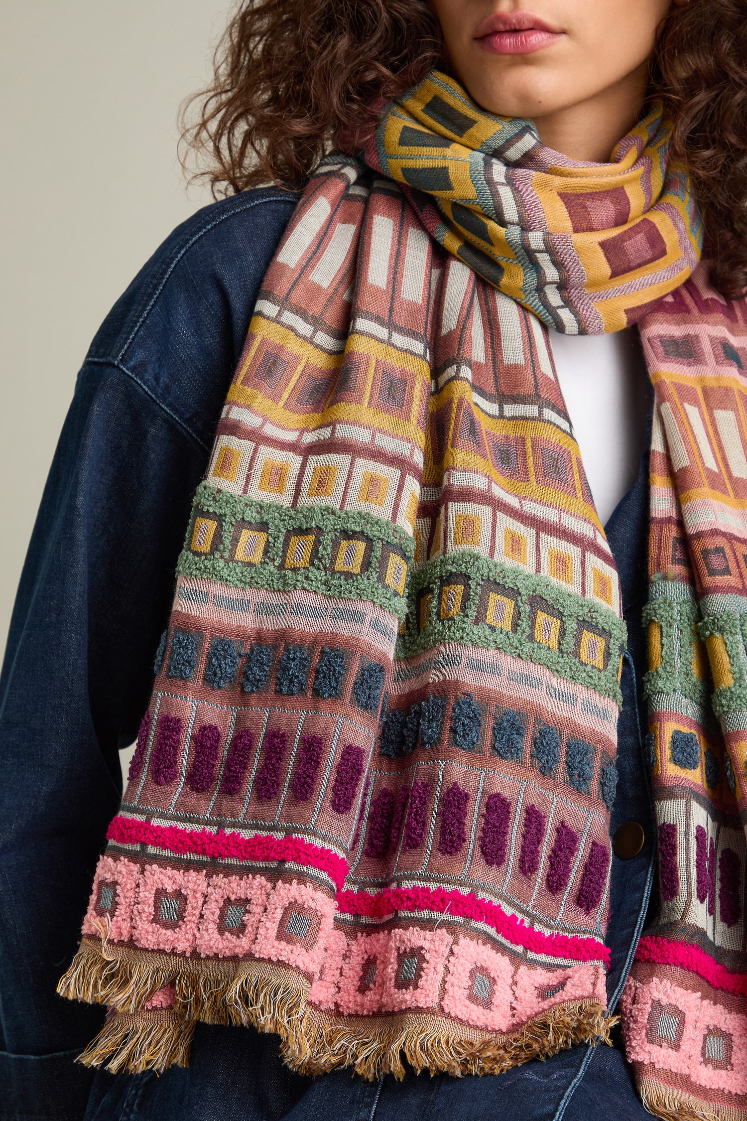 A person wearing the Textured Cotton Scarf, featuring fringe detail and geometric patterns in pink, yellow, green, and maroon, perfectly suited for transitioning between seasons, over a denim jacket.