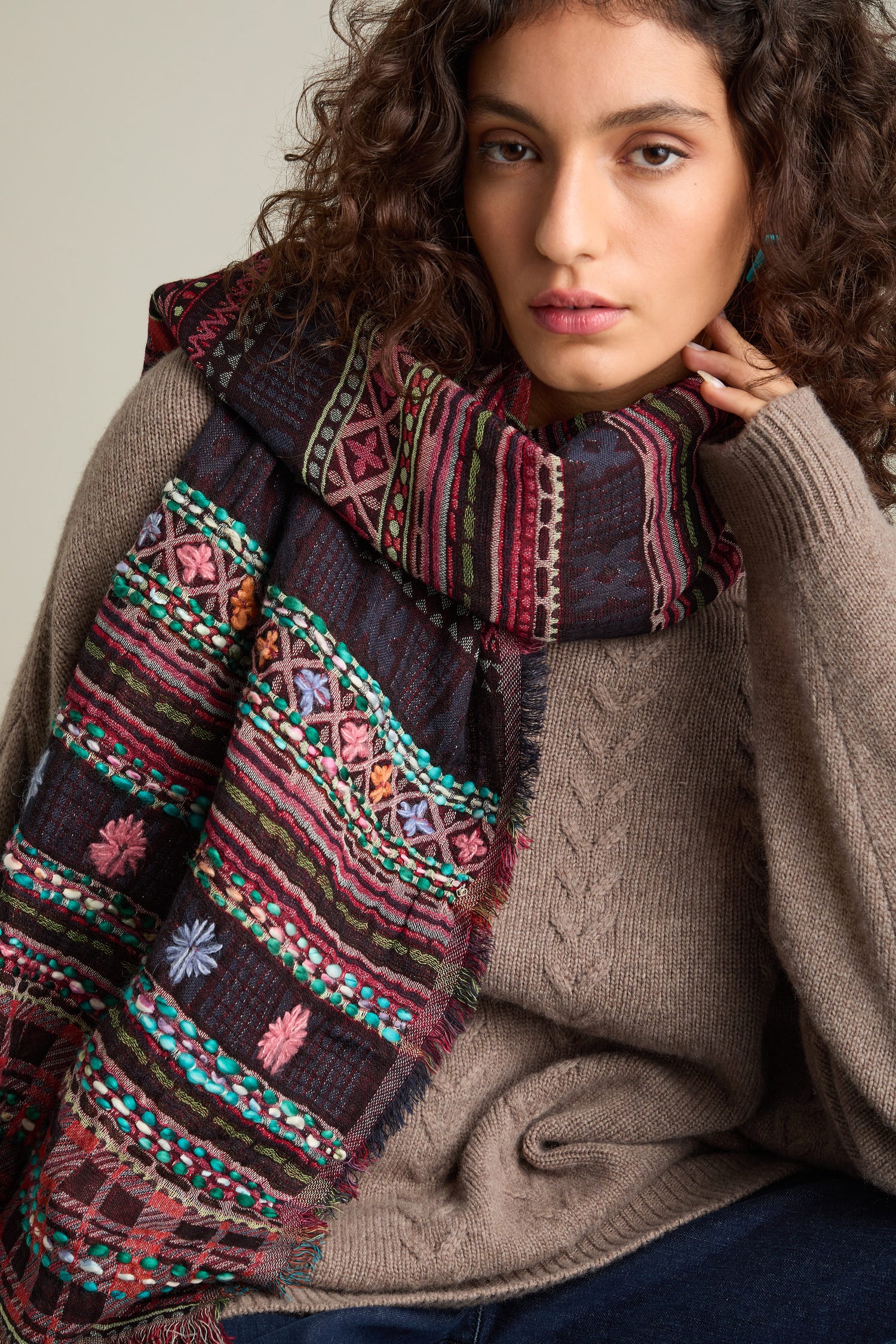 A person with curly hair wears a brown knit sweater and the Hand Embroidered Cotton Scarf, known for its versatility and style, while resting their chin on their hand.